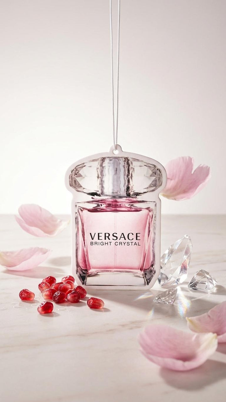 Versace Bright Crystal perfume bottle with a faceted silver cap and pink liquid, shown hanging with a clear cord against a soft light background, surrounded by pink petals, red pomegranate seeds, and clear crystal-like fragments.